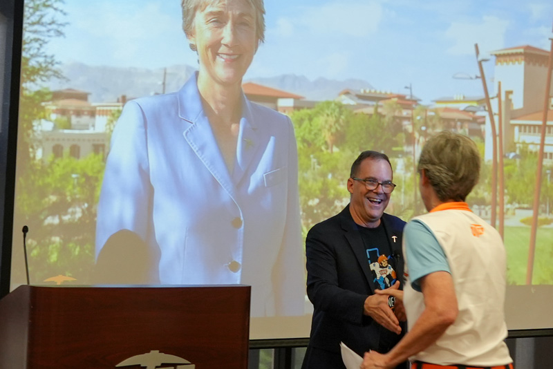 Individual wearing a UTEP Engineering polo shirt and name badge speaks at a podium during a homecoming event, with a presentation slide displaying an alumni profile visible on the screen behind and a microphone positioned beside the speaker.